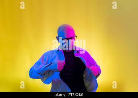 Bearded, bald man with glasses opening his shirt. Isolated on yellow background. Stock Photo