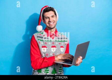 Photo portrait of nice young male hold netbook working remotely wear x-mas print vest hat garment isolated on blue color background Stock Photo