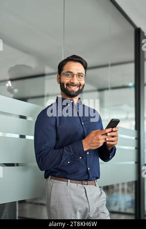 Smiling Indian business man holding phone looking at camera in office. Vertical Stock Photo