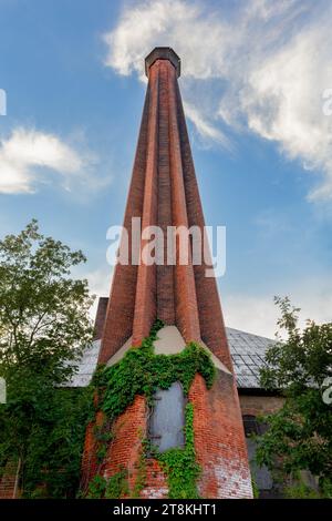 An orange brick smokestack set against a blue sky with an iron door and green ivy at its base.  Concepts could include vintage industry, architecture, Stock Photo