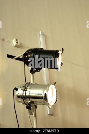 Two turned on theater spotlights on a white wall with wires and an electrical outlet. Close-up. Stock Photo
