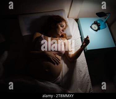 A pregnant woman lies in bed and uses a smartphone. Insomnia. Stock Photo