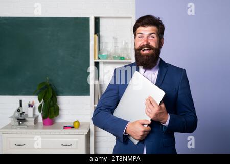 Portrait of funny teacher in classroom. Handsome bearded man near blackboard in classroom. Amazed professor waiting for students, smiling confidently Stock Photo