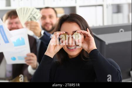 Young happy smiling chinese woman holding two coins of bitcoins in hand instead of glasses rejoices that she had time to buy to produce the moment des Stock Photo