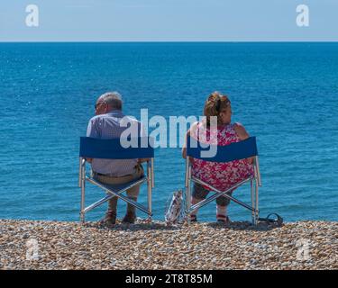 A couple sat on the stone beach in their fold up chairs, facing away from each other on the beach at Lyme Regis, Dorset, England, UK Stock Photo