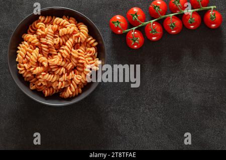 Fusilli pasta, cooked with tomato sauce, branch of cherry tomatoes, on dark background, top view, space to copy text. Stock Photo