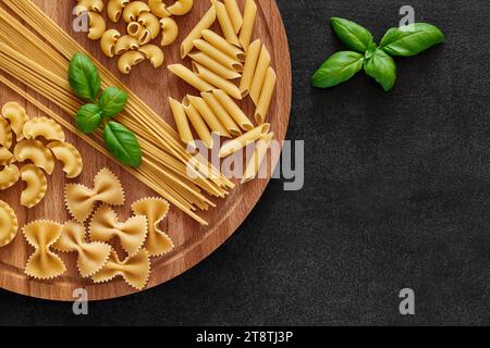 Pasta, set of raw farfalle, spaghetti, pipe, maccheroni, penne, cresta, basil leaf, on round wooden stand, on dark background, top view, space to copy Stock Photo