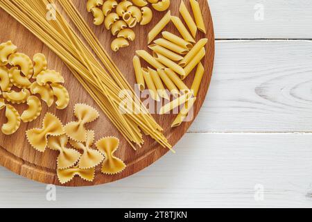 Pasta, mix of raw farfalle, spaghetti, pipe rigatoni, maccheroni, penne rigate, cresta di gallo, on round wooden stand on white board background, top Stock Photo