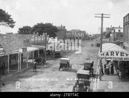 Church Street, Opotiki - Photographed by W J Newell, View looking down Church Street towards Main Street (Elliot Street). In the foreground on the right is Harvey's (fruiter & confectioner). On the lefthandside of the street are various shops including a car hire firm, land agent and butcher. The Post Office building is further down on the left. The Masonic Hotel (with brick facade) can be seen in the far distance on the corner of Church Street and Main Street. Several cars are parked in the street.Photograph taken by W J Newell ca 1920s Stock Photo