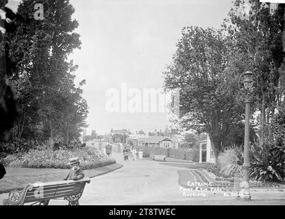 Entrance to Point Erin Park, Auckland, New Zealand, View of the entrance way to Point Erin Park from inside the park. Three people are walking towards the photographer and the man is pushing a fourth person in a wheelchair. In the immediate foreground is a man sitting on a park bench. Houses can be seen in the distance. Taken in early 1900s Stock Photo