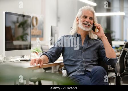 Free lance designer talking on smart phone while working on computer Stock Photo