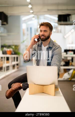Male entrepreneur sitting on table while talking on smart phone Stock Photo