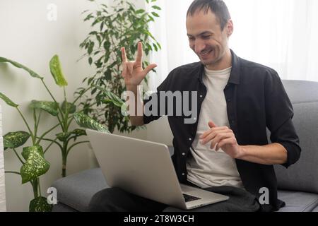 Deaf or hard hearing happy smiling young caucasian man uses sign language while video call using laptop while sitting on the couch at home. Stock Photo