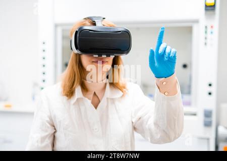 Scientist in VR glasses moves finger imitating touch. Curious blonde woman in rubber gloves tries virtual reality in modern laboratory Stock Photo