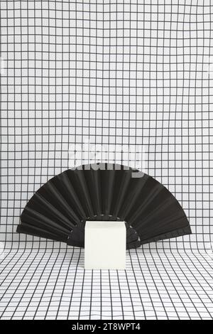 a fan placed behind a white cube as a metaphor for a vampire's cape collar on a black and white grid background. empty object display stand. Minimal s Stock Photo