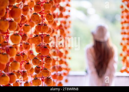 Young woman traveler enjoying dried persimmon hanged on strings to dry a common sight in Da Lat, Vietnam. By hanging them on a rig and placed in an ai Stock Photo