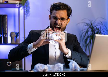 Angry furious Indian man working at office throwing crumpled paper, having nervous breakdown at work, migraine headache, stress management, mental distress problems, losing temper, reaction on failure Stock Photo