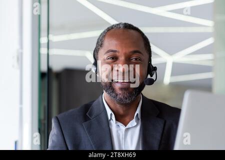 Close-up portrait of a senior smiling African-American man working in an office using a laptop in a headset and smiling at the camera. Stock Photo
