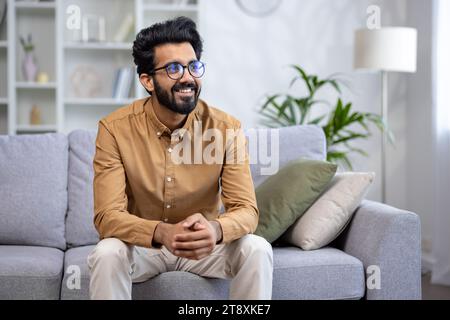 Successful young Indian businessman, freelancer sitting on sofa at home smiling and looking away. Stock Photo