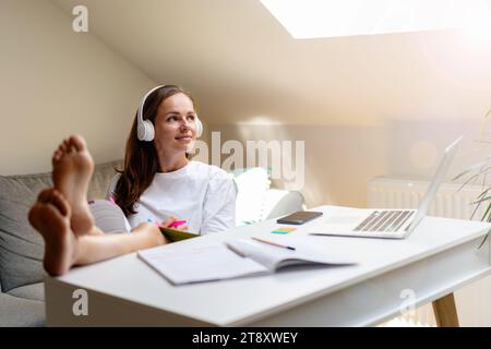 Student's domestic lifestyle. A young woman in white headphones sits with her feet on the table. Stock Photo