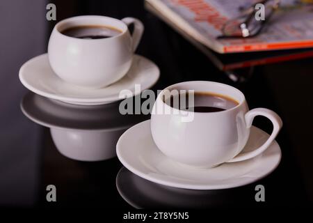 Cups of coffee in white cups and saucers with a magazine in the background. Stock Photo