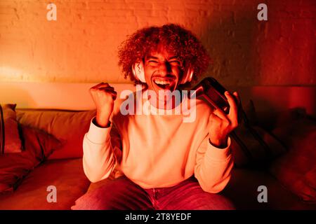 Young man with white headphones playing video games on a sofa in a living room under a red light. Happy young man celebrating a winning Stock Photo
