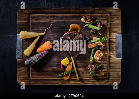 Barbecued meat and fresh vegetables on wooden cutting board, top view and black background Stock Photo