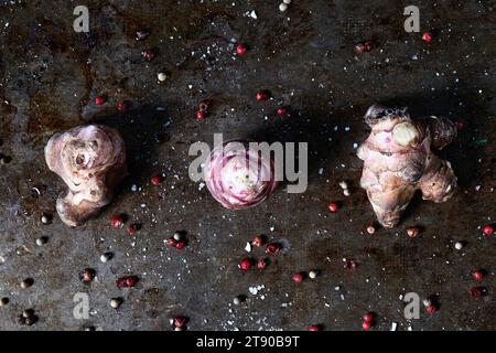 Three top-view fresh Jerusalem artichokes laid horizontally on a baking tray with herbs and coarse salt Stock Photo