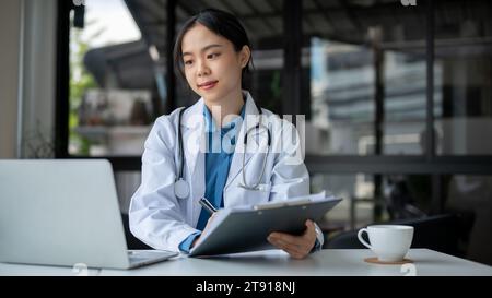 A beautiful and experienced young Asian female doctor in a white gown is working at her desk in the hospital, reading medical reports online. Stock Photo