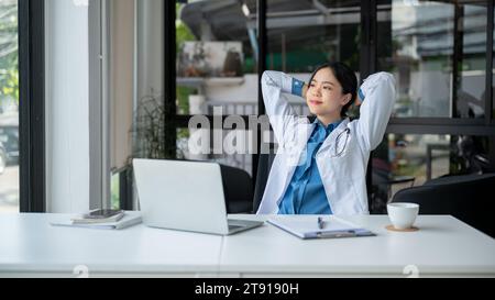 A relaxed and carefree Asian female doctor is putting her hands behind her head, looking outside, daydreaming and feeling fresh while taking a break a Stock Photo