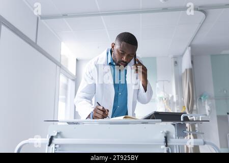 African american male doctor talking on smartphone and taking notes in hospital room Stock Photo