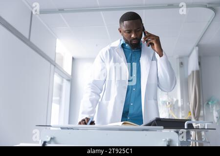 African american male doctor talking on smartphone and taking notes in hospital room Stock Photo