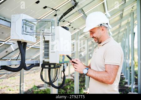 Enthusiastic man happy about launching solar panel. Smiling worker reporting on well done job. Man in helmet texting in his phone on background of PV battery. Stock Photo