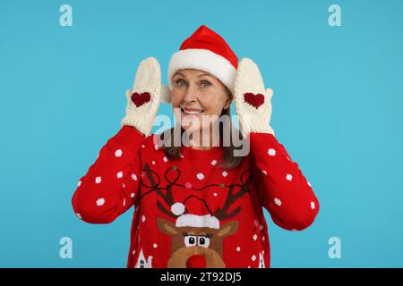 Senior woman in Christmas sweater, Santa hat and knitted mittens on light blue background Stock Photo