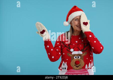 Senior woman in Christmas sweater, Santa hat and knitted mittens on light blue background. Space for text Stock Photo