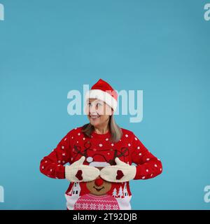 Happy senior woman in Christmas sweater, Santa hat and knitted mittens on light blue background Stock Photo
