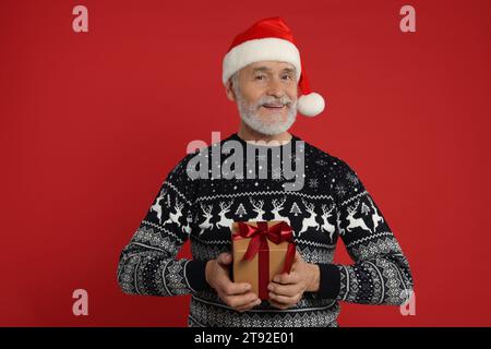 Senior man in Christmas sweater and Santa hat holding gift on red background Stock Photo