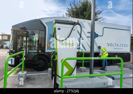 A Volta Zero truck is parked at an Allego charging station. A man in reflective jacket inspects it with a tree and clear blue sky in the background Stock Photo