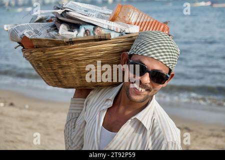 A young, smiling Indian snack vendor wearing sunglasses and carrying a basket with his his wares; at Chowpatty Beach, Girgaum, Mumbai, India Stock Photo