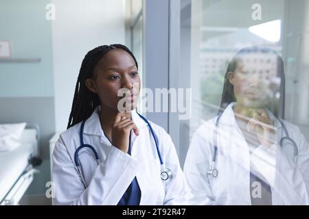 Thoughtful african american female doctor wearing lab coat looking through window in hospital room. Medicine, healthcare, work and hospital, unaltered Stock Photo