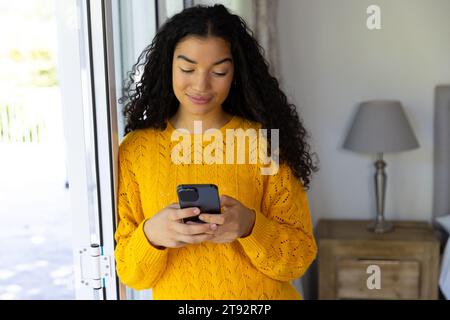 Happy biracial woman in yellow sweater using smartphone at home Stock Photo