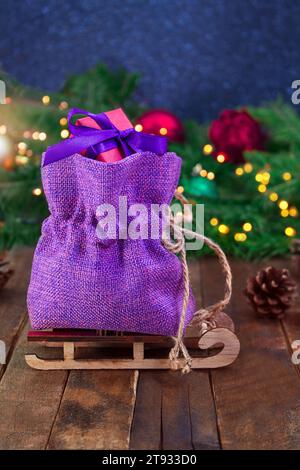 Canvas purple bag with red gift box on sled on wooden boards. Spruce branches, toys and defocused lights. Christmas, New Year, sale. Soft focus. Eveni Stock Photo