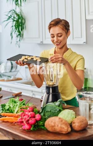 pleased vegetarian woman putting sliced banana in electric blender near fresh vegetables in kitchen Stock Photo