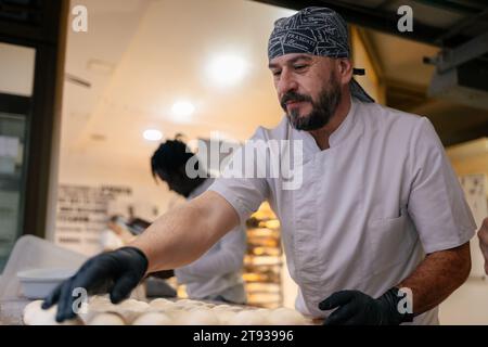 Adult baker dressed in white clothes and a black cap making balls of dough and placing them on a tray in a bakery Stock Photo