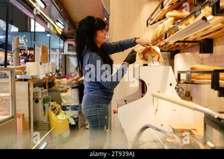 Adult woman with black hair and blue clothes putting a loaf of bread in a bag after slicing it for a customer in a bakery Stock Photo