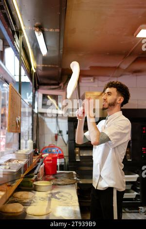 Portrait of young arabic pizza man in white apron throwing the dough in the air in a pizzeria Stock Photo