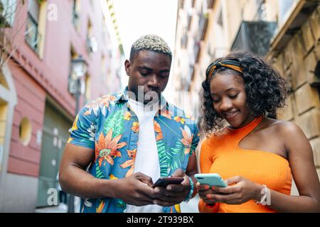 Young black couple in colorful and casual clothes walking around the city while using their smartphone in the street Stock Photo