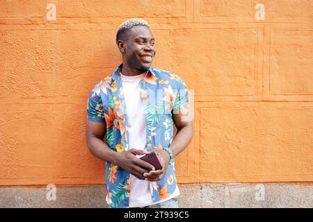 Portrait of a handsome young black man wearing casual clothes looking to his left smiling while holding his smartphone leaning against an orange wall Stock Photo