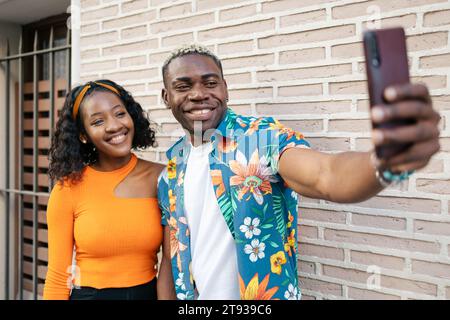 Young black couple wearing casual and colorful clothes taking a selfie and smiling while they walk around the city Stock Photo
