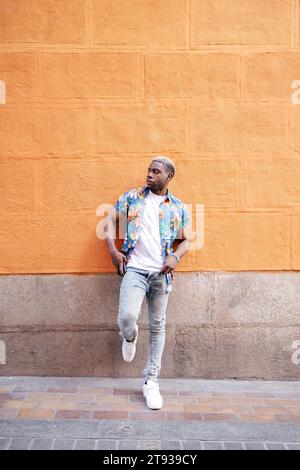 Portrait of a handsome young black man wearing casual clothes keeping his smartphone in the pocket leaning against an orange wall Stock Photo